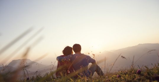 A couple sits on a hill overlooking the mountains at sunset with their partner.