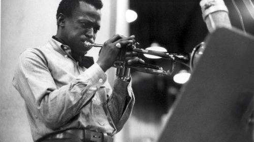 A black and white photo of a man playing a trumpet in a tucked-in shirt.