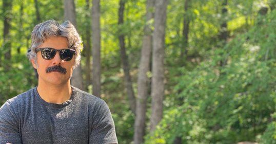 A man with sunglasses standing in a wooded area.