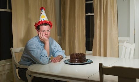 A man with a birthday cake on his head sits at a table.