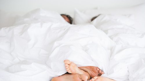 Two people's feet under a white blanket in bed, hinting at their relationship and sleeping habits.