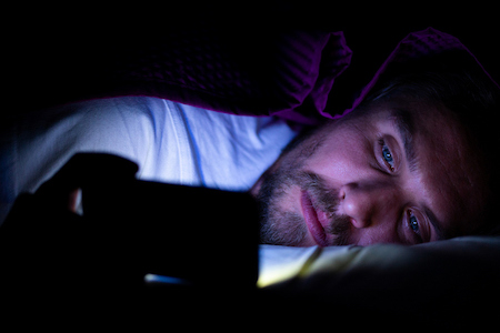 A man laying in bed with a cell phone, browsing the internet.