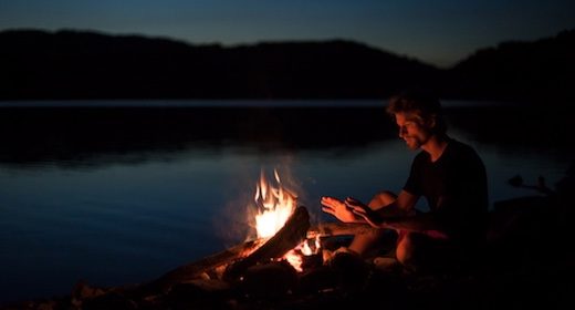 A man sitting by a campfire near a lake at night, experiencing the rewild of nature.
