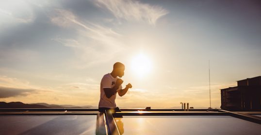 A man stands on top of a roof at sunset, casting a long shadow.