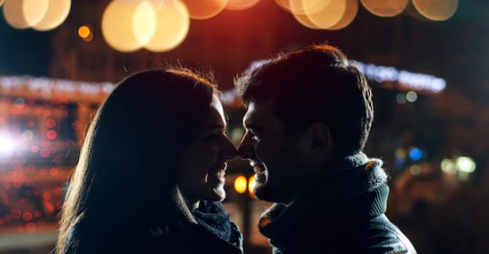 A couple gazing at each other late at night in a bustling city.