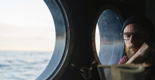 A bearded man gazes out the window of a ship.