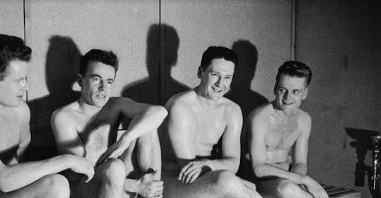 A group of naked men observing sauna etiquette while sitting on a bench.