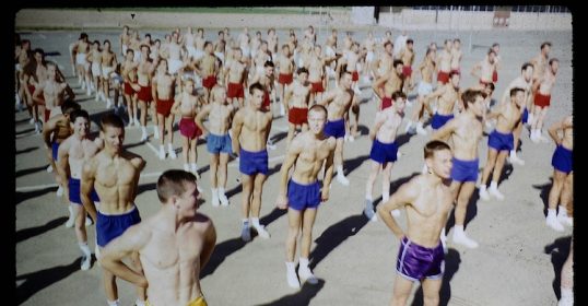 A group of men in swim trunks standing in a line participate in History's Hardest P.E. Program.