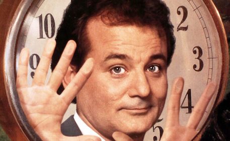 A man in a suit is holding his hands up in front of a clock, possibly reflecting on his Podcast or Groundhog Day.