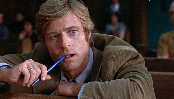 A man wearing corduroy sits in a courtroom with a pen in his hand.