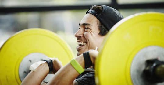 A person smiling while lifting a barbell with yellow weights at the gym and listening to a 