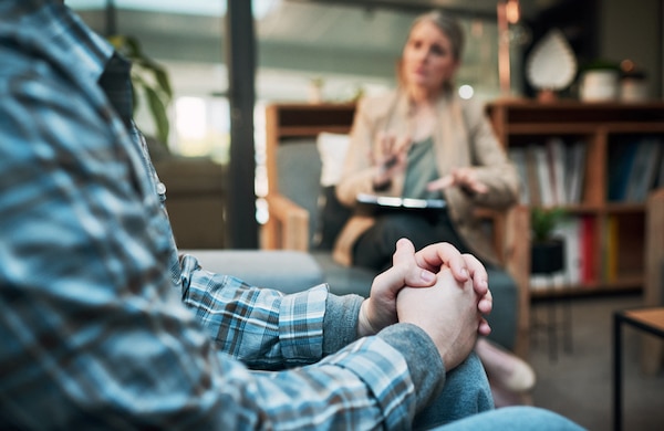 Man in a plaid shirt sitting and listening intently to a woman therapist who is taking notes during a psychology counseling session in a cozy office environment.