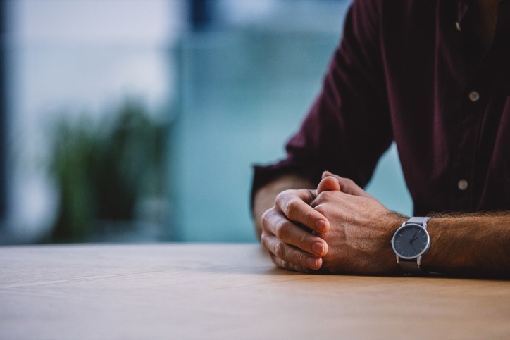 Close-up of a person's clasped hands resting on a table, waiting, wearing a watch on the left wrist.