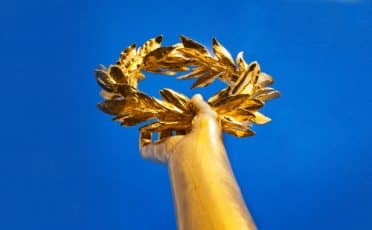 Golden laurel wreath, symbolizing attributes of greatness, held aloft against a blue sky.