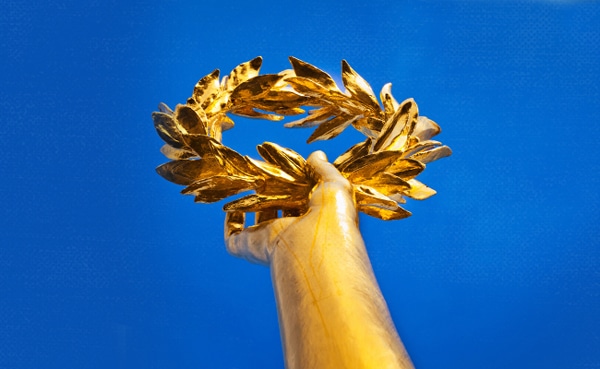Golden laurel wreath, symbolizing attributes of greatness, held aloft against a blue sky.