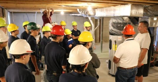 Group of construction trainees in hard hats receiving Trades School instruction in a building's basement.