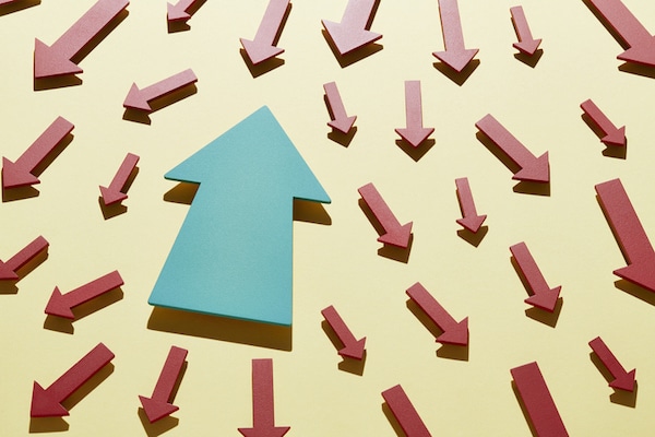 A blue arrow pointing upwards amidst numerous red arrows pointing downwards against a yellow background, symbolizing anti-mimetic life choices.