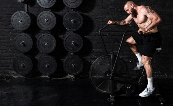 A man with tattoos working out on an assault bike in a gym with weights in the background.