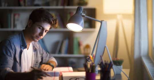 A young man studying at a desk under a lamp at night, gaining knowledge.
