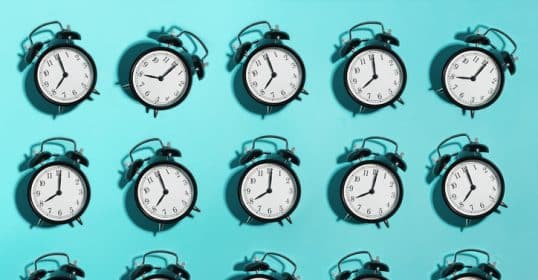 An array of identical black alarm clocks against a teal background, each showing a different time, symbolizes the real reason behind procrastination discussed in Podcast #931.