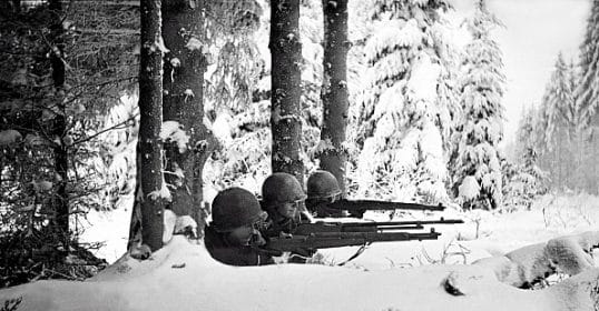 Two soldiers in winter camouflage lie prone with rifles during the Battle of the Bulge, aiming through snowy forest terrain.