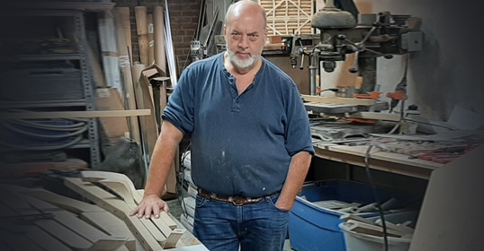 A carpenter standing in a woodworking shop with workbenches and tools in the background.
