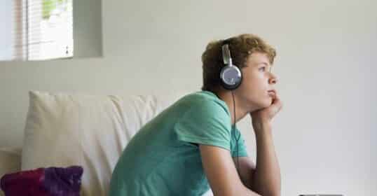 A teenage son sitting on a bed wearing headphones, looking thoughtfully out of a window.