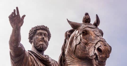 Bronze statue of a stoic emperor mounted figure with raised hand under a cloudy sky.