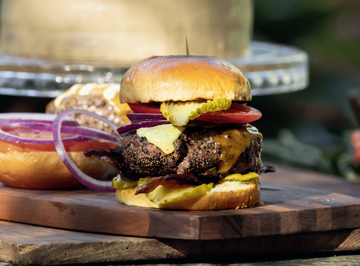 A gourmet cheeseburger with pickles, tomatoes, red onions, and lettuce served on a wooden board as part of Grandaddy's Sunday Dinner.