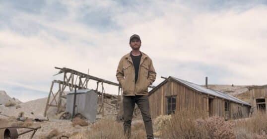 Man standing confidently in front of an old mining structure in a ghost town, wearing a jacket and cap.