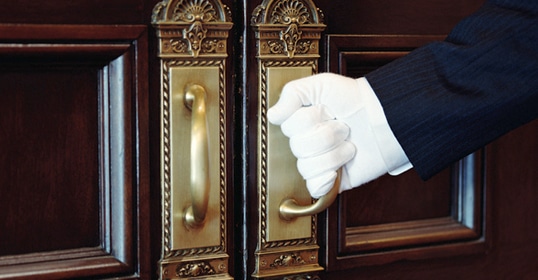 Person in a suit with white gloves, serving as a butler, opening an ornate wooden door.