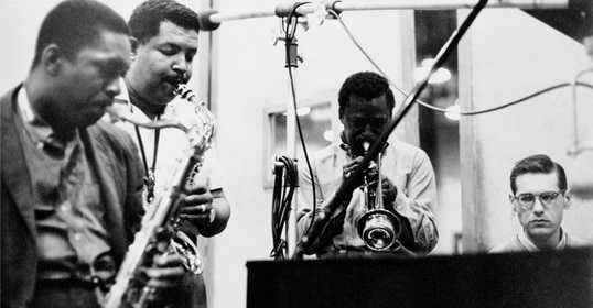 Three men playing saxophone and trumpet in a recording studio, observed by another man wearing glasses. This black and white photo captures the essence of the 