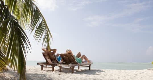 Two people relaxing on sun loungers by the beach under clear skies during their vacation.