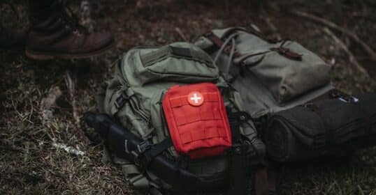 A red first aid kit, essential for grid-down medicine, attached to a green backpack with hiking boots and other camping gear on a forest floor.
