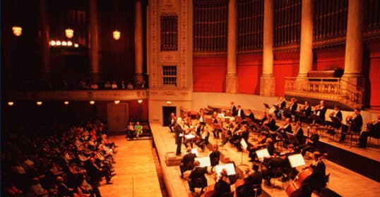 Orchestra performing classical music on stage in a grand hall with an audience in the foreground.