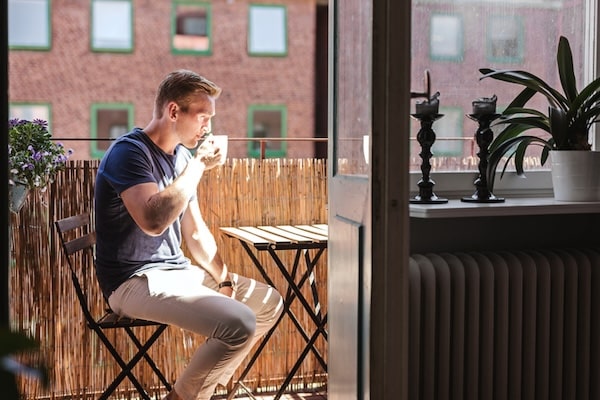 A man sitting at a small outdoor table embraces his everyday rituals by drinking from a cup on a sunny balcony, with green plants and a decorative item visible inside.