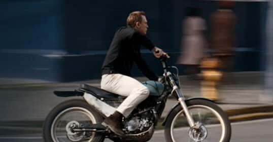 A man in white jeans and casual attire riding a motorcycle swiftly on an urban street, with motion blur indicating speed.