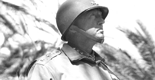 A person in military uniform and helmet, reminiscent of Patton, gazes to the right against a backdrop of trees.