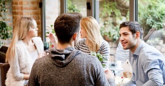 Four people sit at a table in a café, engaged in conversation. Two men, showcasing the nuances of male friendships, face the camera, while two women are seen in profile. The background reveals a bright, greenery-filled outdoor area.