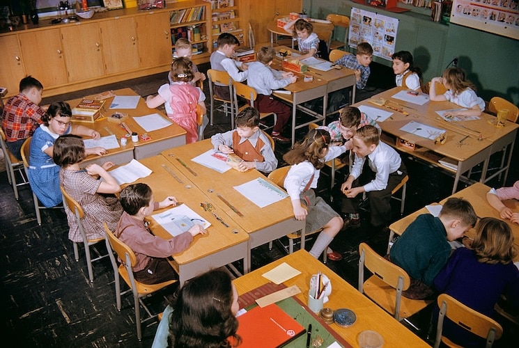 Children are seated at tables in a classroom, engaging in various activities and conversations with each other. Some are drawing on papers while others discuss their thoughts or look around. Bookshelves filled with resources on moral education stand proudly in the background.