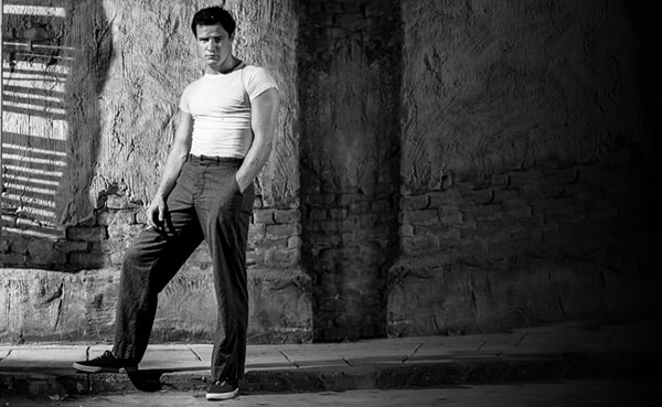 A man in a fitted, tucked-in white t-shirt and dark pants stands casually against a textured wall with one hand in his pocket, in a black-and-white photo.