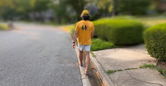 A person wearing a yellow shirt and white shorts enjoys a morning walk, barefoot along a suburban street, near the curb and a grassy area.