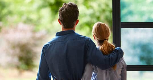 A man and a woman stand together by a window, facing outside. The man has his arm around the woman, embodying essential relationship tips for a stronger bond. Both dressed casually, they look out at the greenery visible through the window.