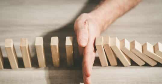 A hand stops falling wooden dominoes on a table, halting a chain reaction, much like how a differentiated self can manage group anxiety.