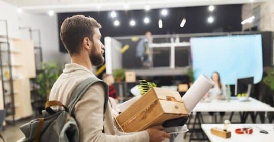 A man carrying personal items in a box and backpack walks through an office space with multiple desks and busy coworkers in the background, as he prepares to break up with his job to pursue a new career.