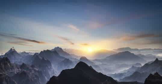 A panoramic view of a mountainous landscape at sunrise, with layers of rugged peaks and ridges extending into the distance under a clear sky, as if each mountain represents a step in personal development.