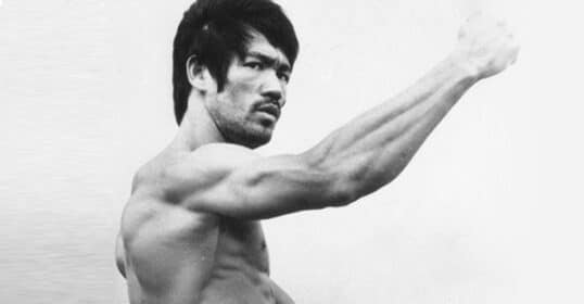 A shirtless man with a muscular build, reminiscent of Bruce Lee, strikes a pose with his right arm extended in a punching motion and his left arm bent. He has short, dark hair and a serious expression, showcasing the results of an intense forearm workout.