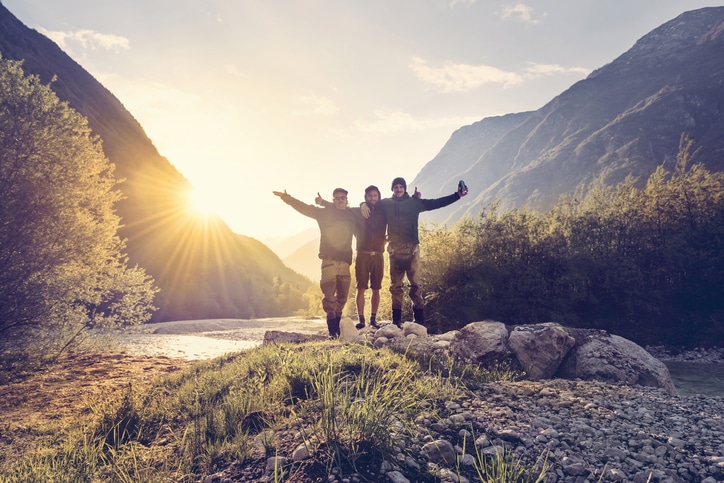 Three people stand together with arms raised in celebration on a rocky riverbank, surrounded by lush mountains, with the sun setting behind them—an ideal scene of enduring friendships.