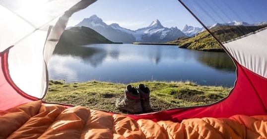 View from inside a tent showcasing a pair of hiking boots outside, along with essential outdoor gear, against the backdrop of a tranquil lake, grassy shores, and snow-capped mountains under a clear blue sky.