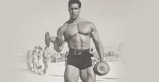 A man performs a bicep curl with a dumbbell on a beach while wearing shorts and a weightlifting belt, showcasing his dedication to fitness. Several people are in the background.